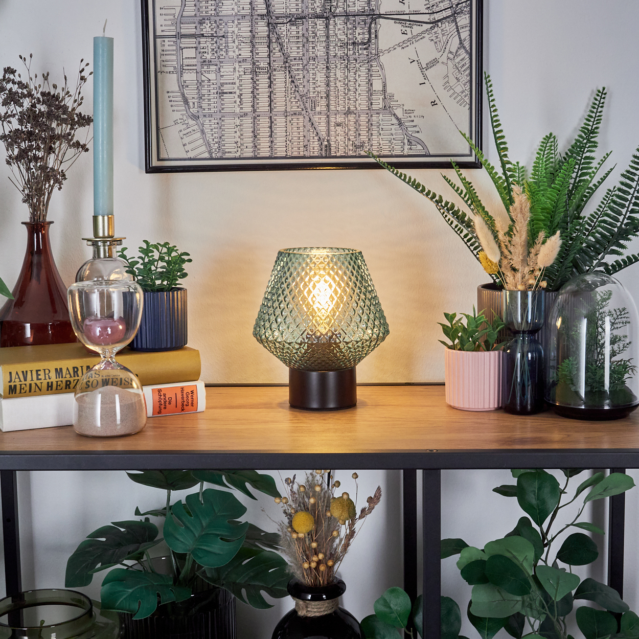 Eine Lampe mit grünem Schirm steht auf einem Holzregal mit Pflanzen, Büchern und einer Karte. Kerze und Sanduhr sind sichtbar.