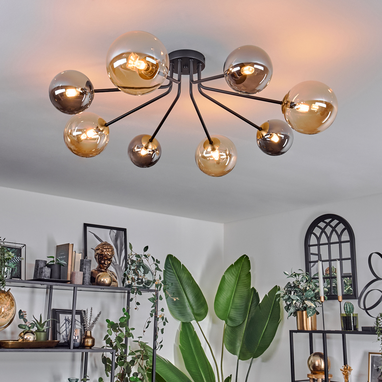 Eine moderne Deckenleuchte mit kugelförmigen Glasschirmen hängt über einem Raum mit Pflanzen und Regalen. Der Raum ist gut beleuchtet und stilvoll.