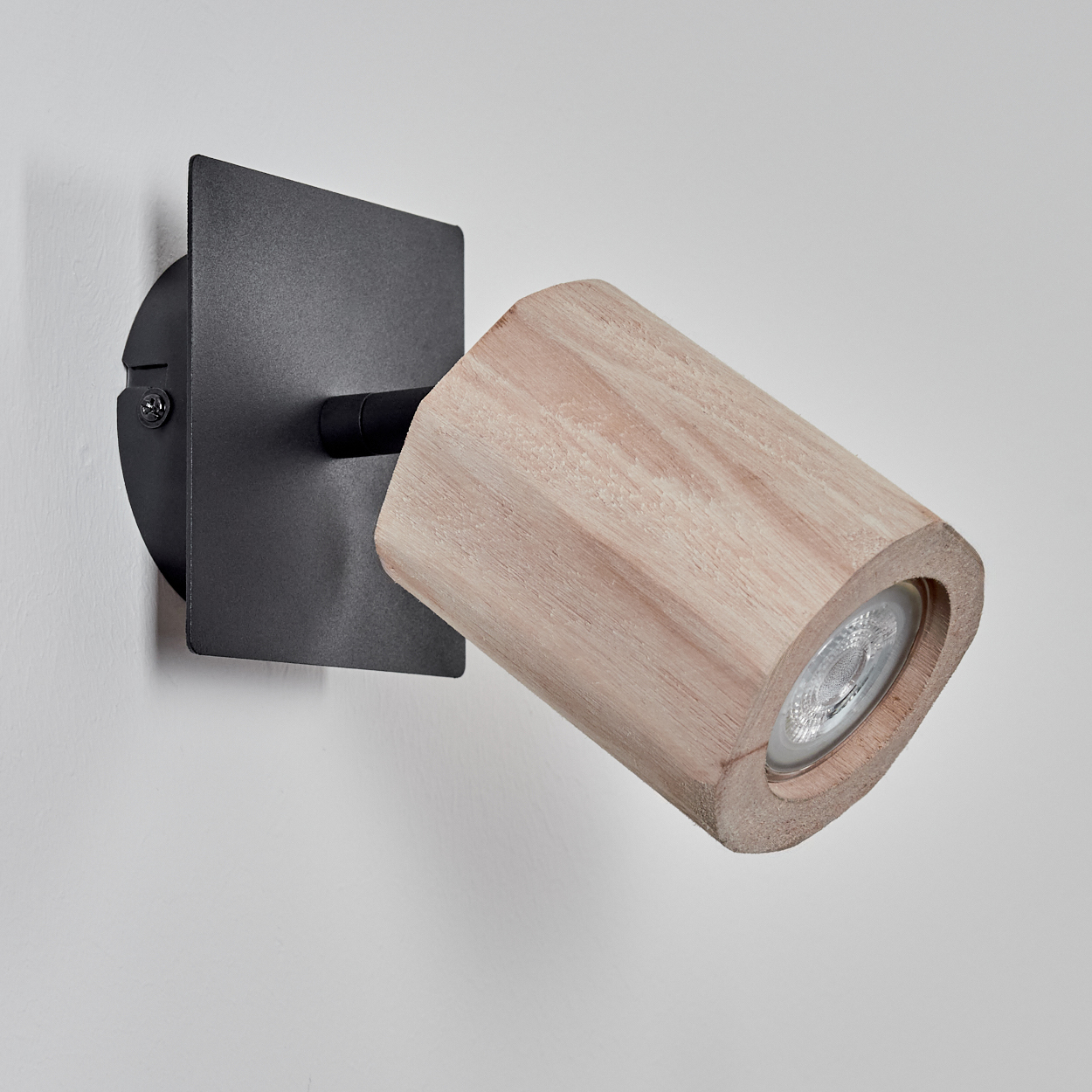 Ein Holzstrahler ist auf einer schwarzen quadratischen Platte montiert, die an einer weißen Wand befestigt ist. Das Licht ist nach außen gerichtet.
