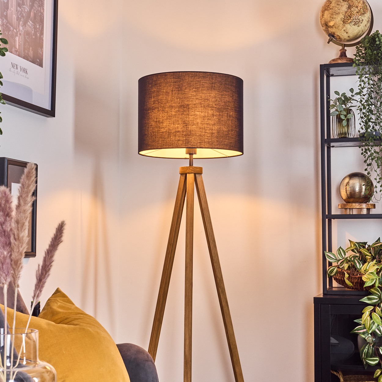 Eine Holz-Stativlampe mit schwarzem Lampenschirm. Ein Regal mit Pflanzen und einem Globus. Auch ein Sofa mit gelben Kissen ist zu sehen.