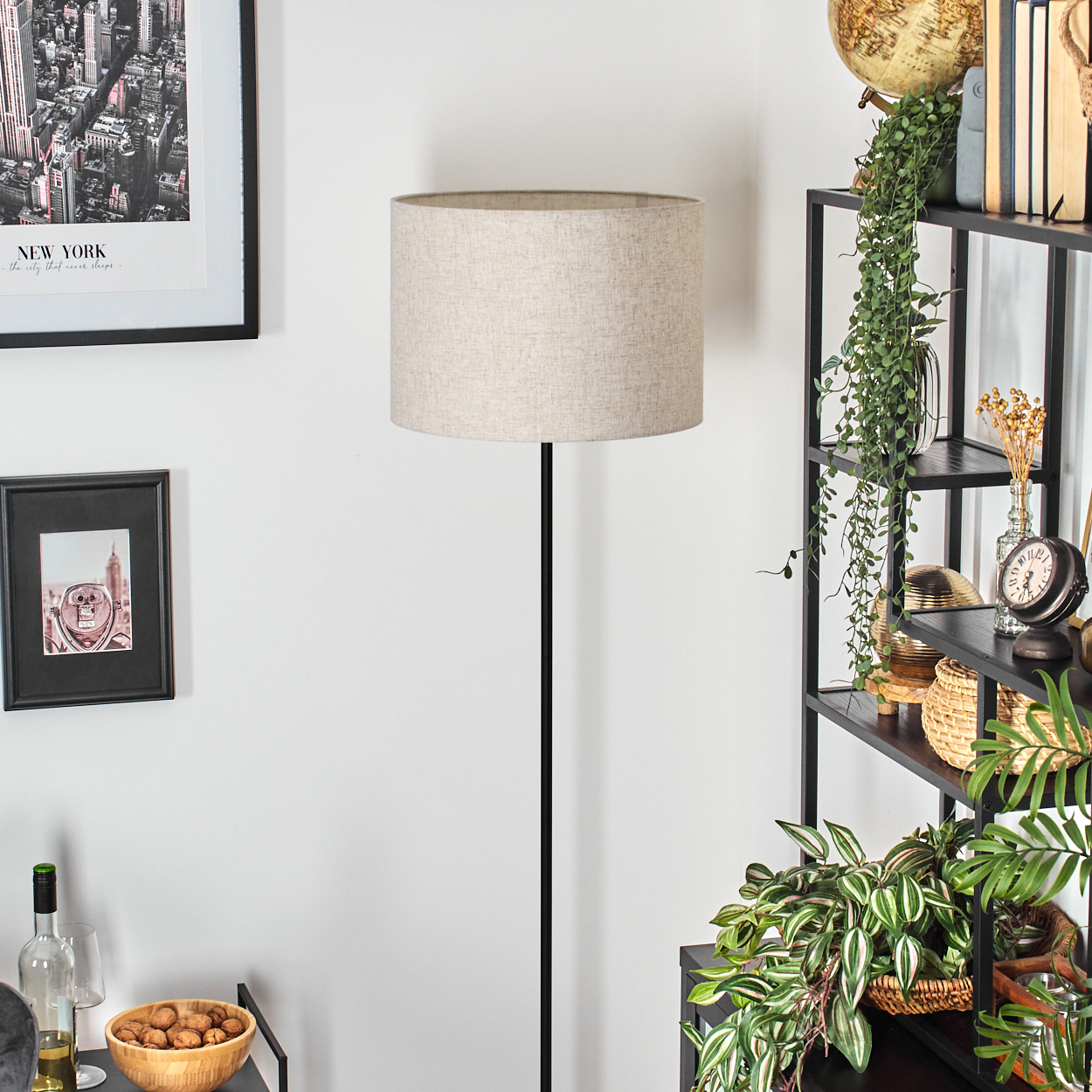 Ein Raum mit einer Stehlampe. Ein Regal voller Dekoration befindet sich auf der rechten Seite. Bilder hängen an der Wand.