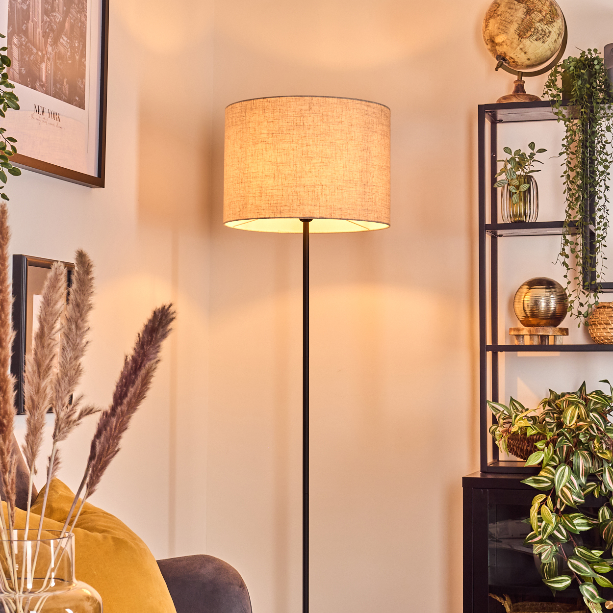 Eine Stehlampe steht in einer Ecke. Rechts ein Regal mit Deko. Links ein Bild mit getrockneten Pflanzen in einer Vase.