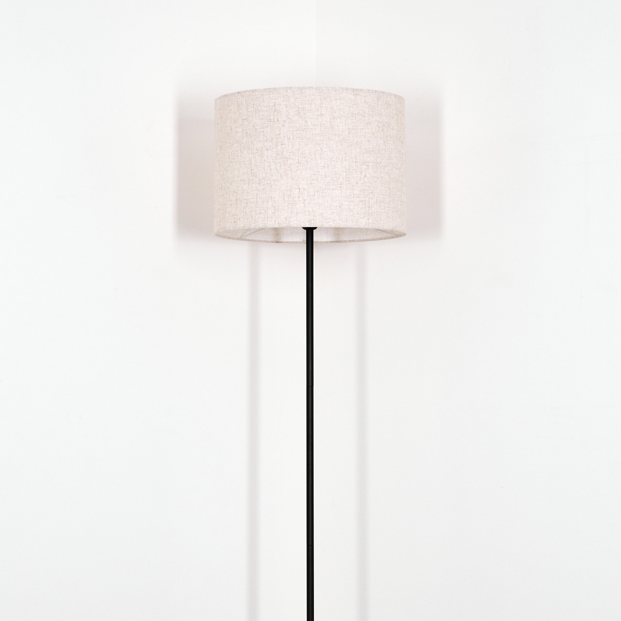 Ein beiger Lampenschirm sitzt auf einer schwarzen Stange. Die Lampe steht vor einer weißen Wand. Schatten werden auf die Wand geworfen.