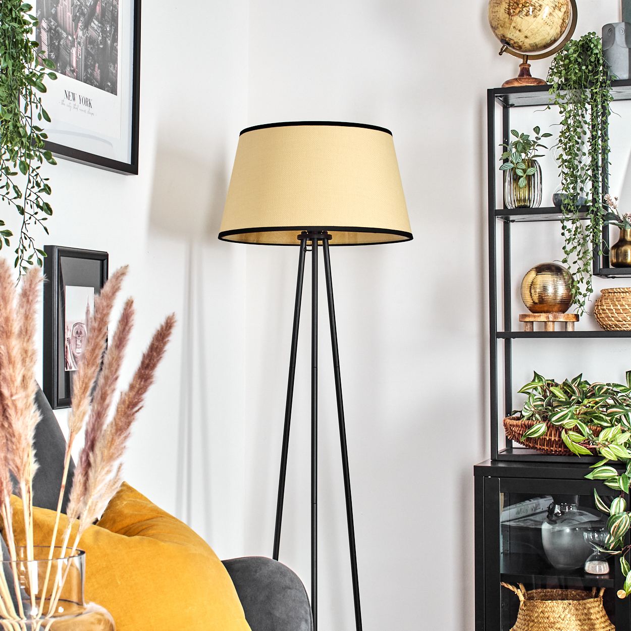 Eine beige Lampe steht in einer Ecke. Ein schwarzes Regal mit Pflanzen und einem Globus ist sichtbar. Links ein gelbes Kissen.