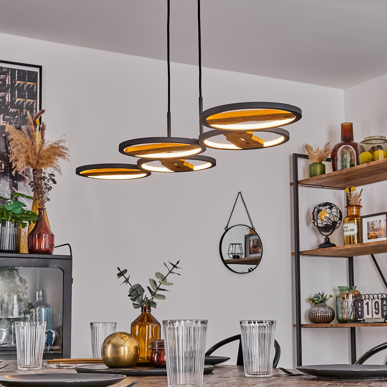 Esszimmer mit Holztisch. Moderne kreisförmige Lampen hängen darüber. Dekorationen umfassen Vasen, Spiegel und Regale.