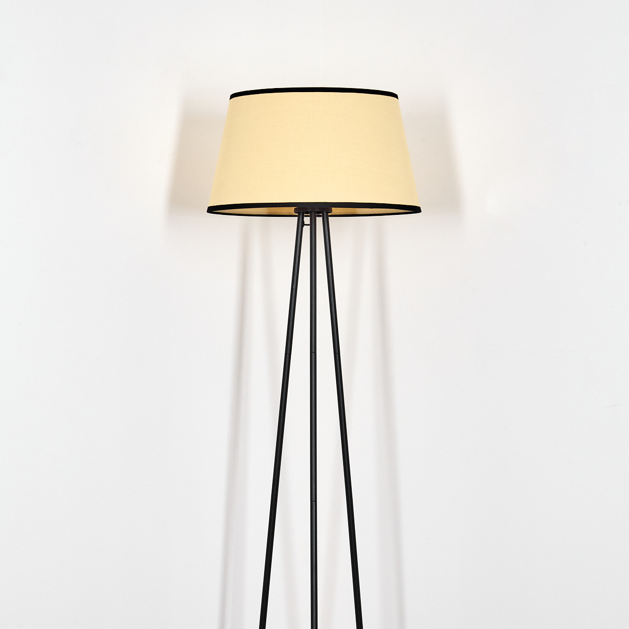 Eine Stehlampe mit beigem Lampenschirm und schwarzem Rand. Die Lampe hat einen schwarzen Dreibein-Sockel vor einer weißen Wand.