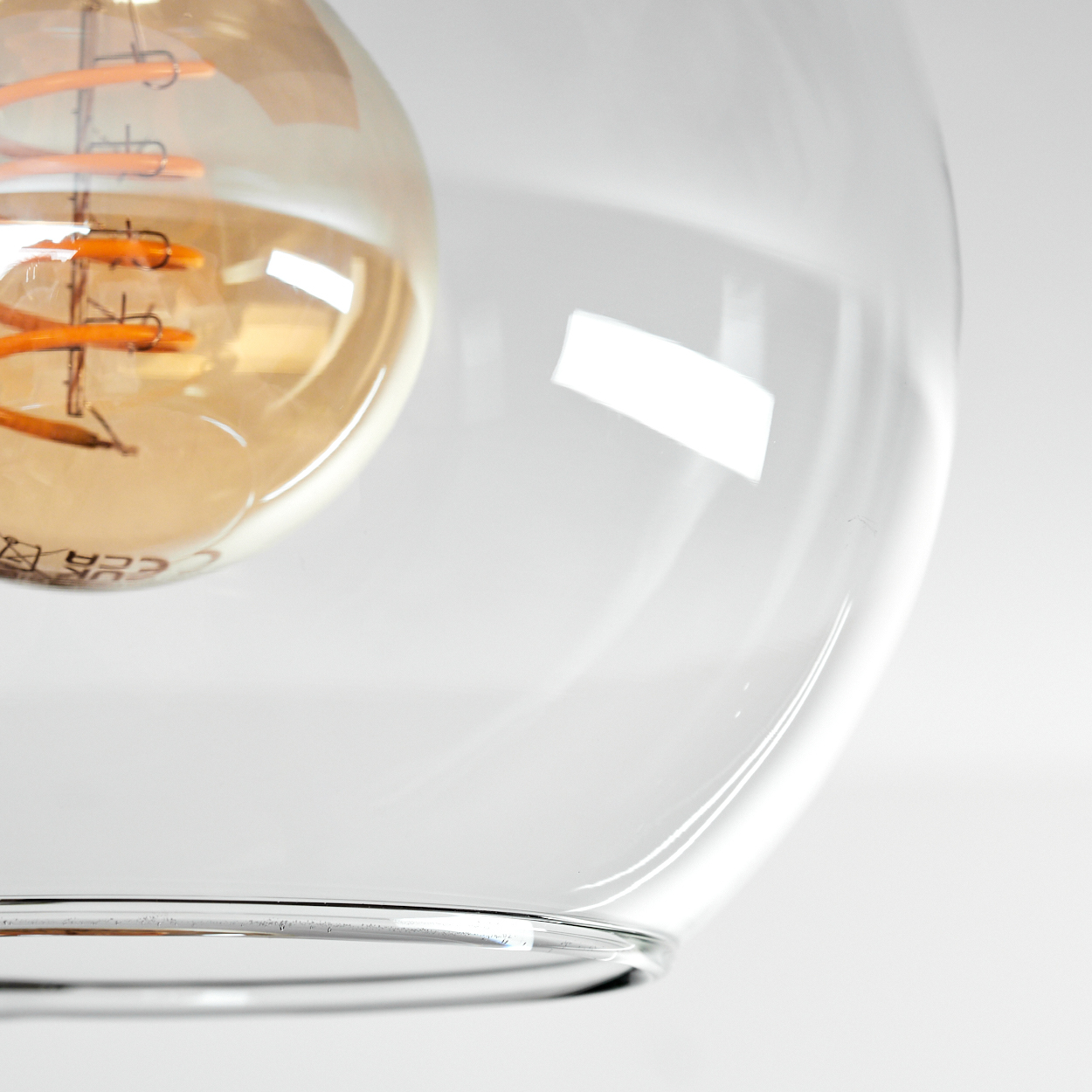 Nahaufnahme einer klaren Glas-Pendelleuchte mit warmtoniger Glühbirne. Der Hintergrund ist weiß.