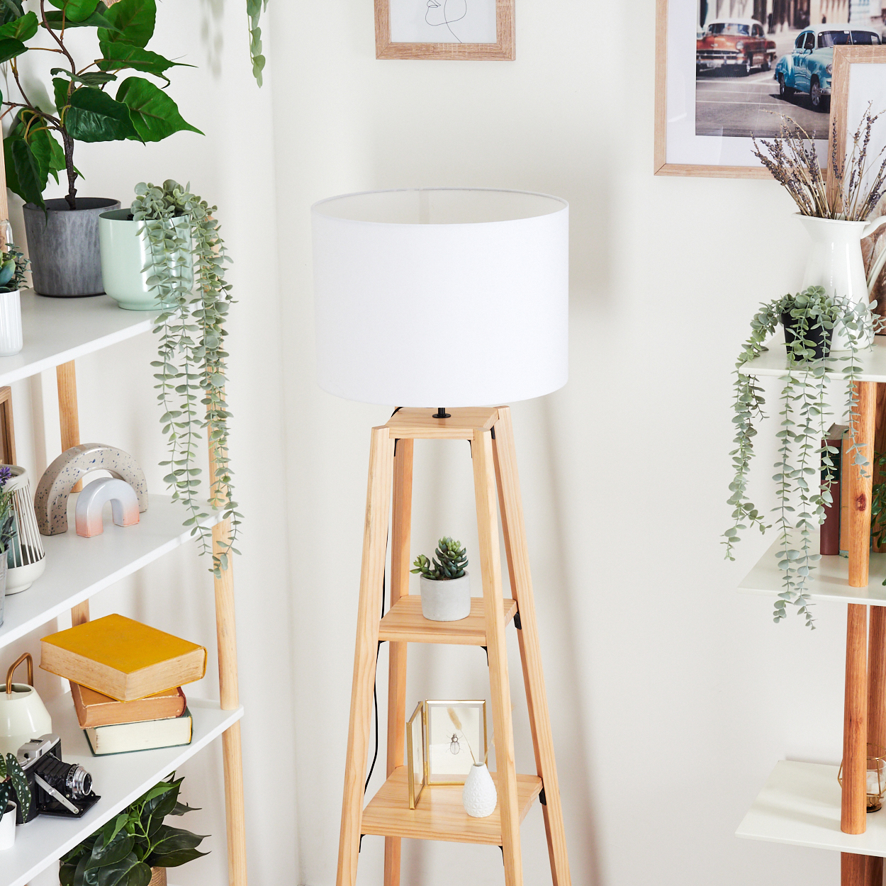 Eine Holzlampe mit weißem Schirm steht neben Regalen mit Pflanzen, Büchern und Dekorationsartikeln. Kunstwerke hängen an der Wand.
