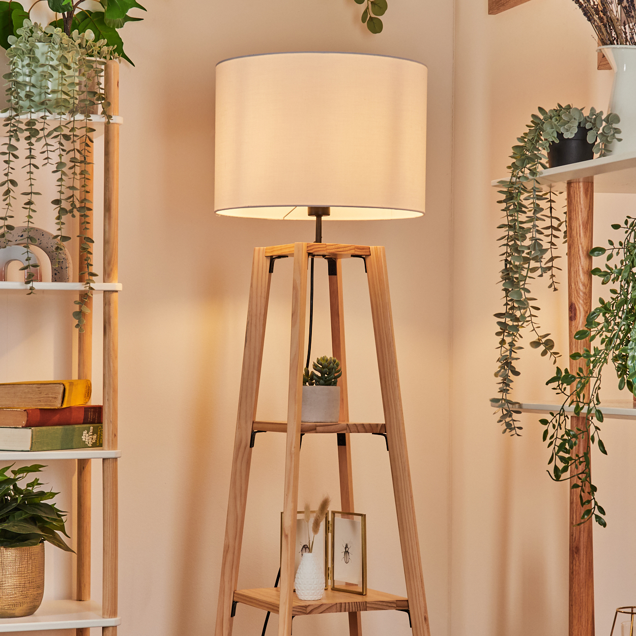 Eine Holzlampe mit weißem Lampenschirm steht in einem Raum. Regale mit Pflanzen und Büchern befinden sich auf beiden Seiten der Lampe.