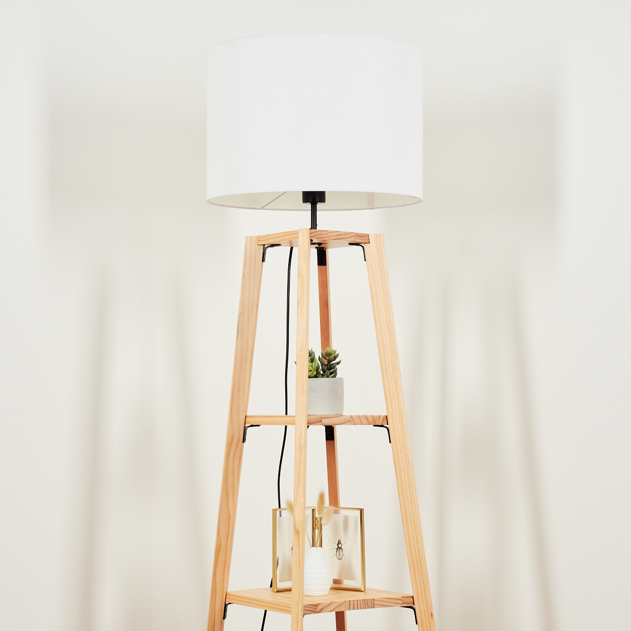 Eine Stehlampe aus Holz mit weißem Lampenschirm. Sie hat Regale mit einer Pflanze, einer Vase und einem gerahmten Bild. Der Hintergrund ist weiß.