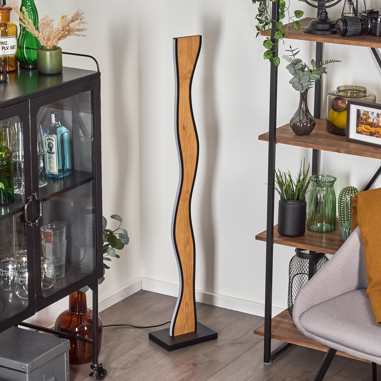 Eine Holzlampe mit wellenförmigem Design steht in einem Raum. Regale und ein Schrank mit Gegenständen befinden sich im Hintergrund.