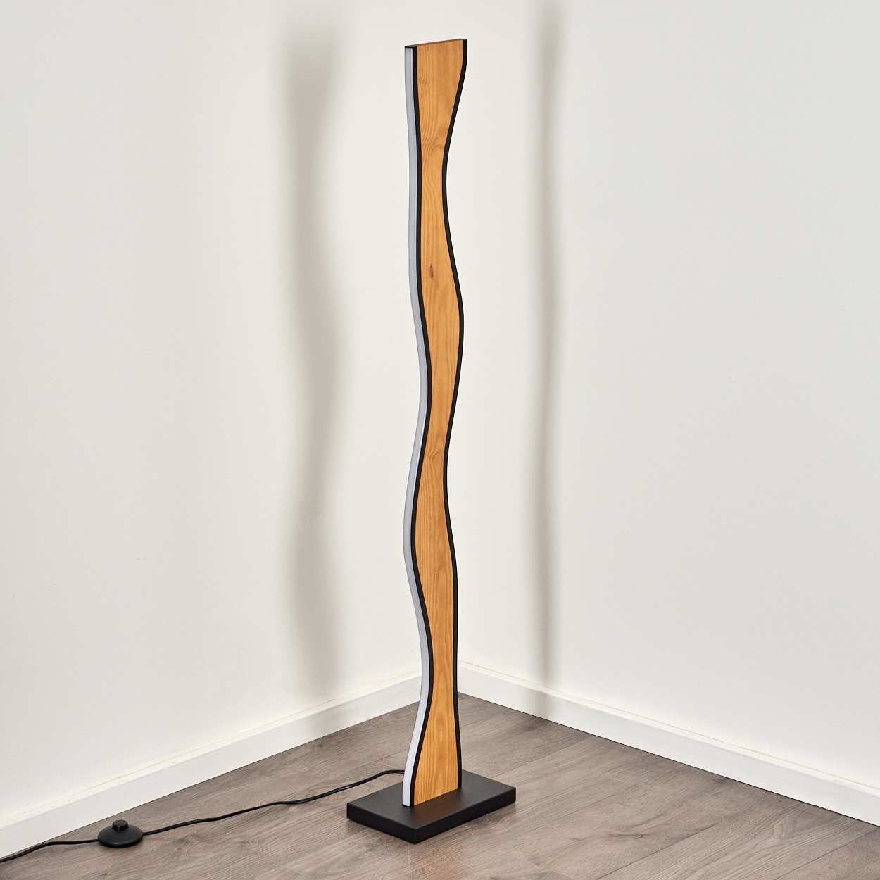 Eine moderne Stehlampe mit geschwungenem Holzdesign steht auf einem schwarzen Sockel. Sie steht vor einer weißen Wand auf einem Holzboden.