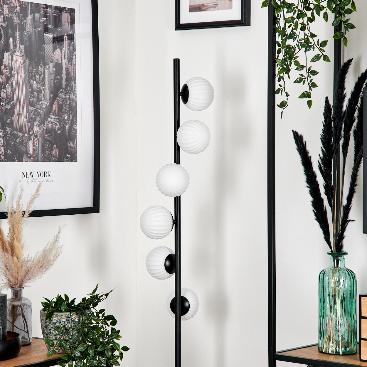Ein Raum mit einer Stehlampe mit weißen Kugelleuchten, einem gerahmten New York-Druck und dekorativen Pflanzen in Vasen.