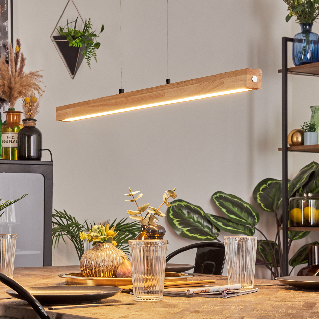 Eine Holzlampe hängt über einem Tisch mit Gläsern und einem Tablett mit Dekorationen. Pflanzen und Regale sind im Hintergrund sichtbar.