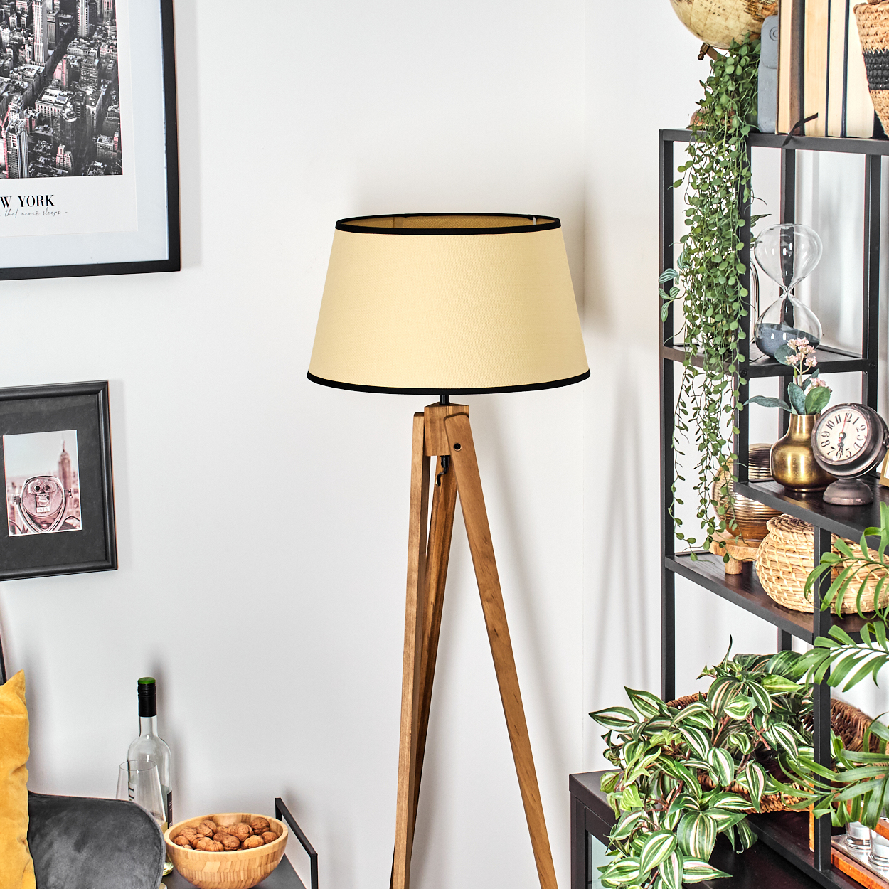 Eine Holz-Stativ-Stehlampe mit beigem Lampenschirm steht neben einem schwarzen Regal mit Pflanzen und Deko.