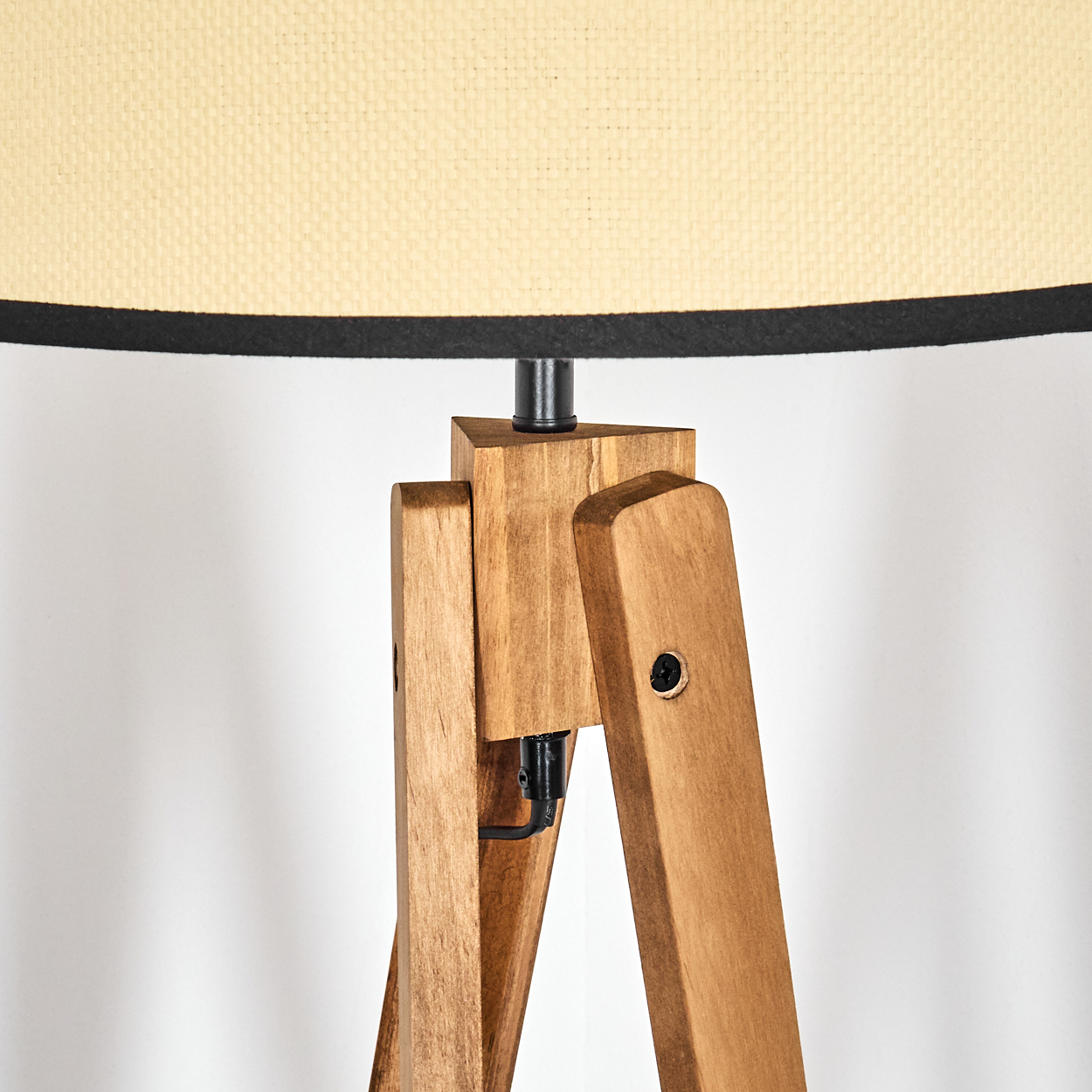 Eine Stehlampe mit Holzstativ. Sie hat einen beigefarbenen Lampenschirm mit schwarzem Rand. Die Beine sind braun, der Hintergrund ist weiß.
