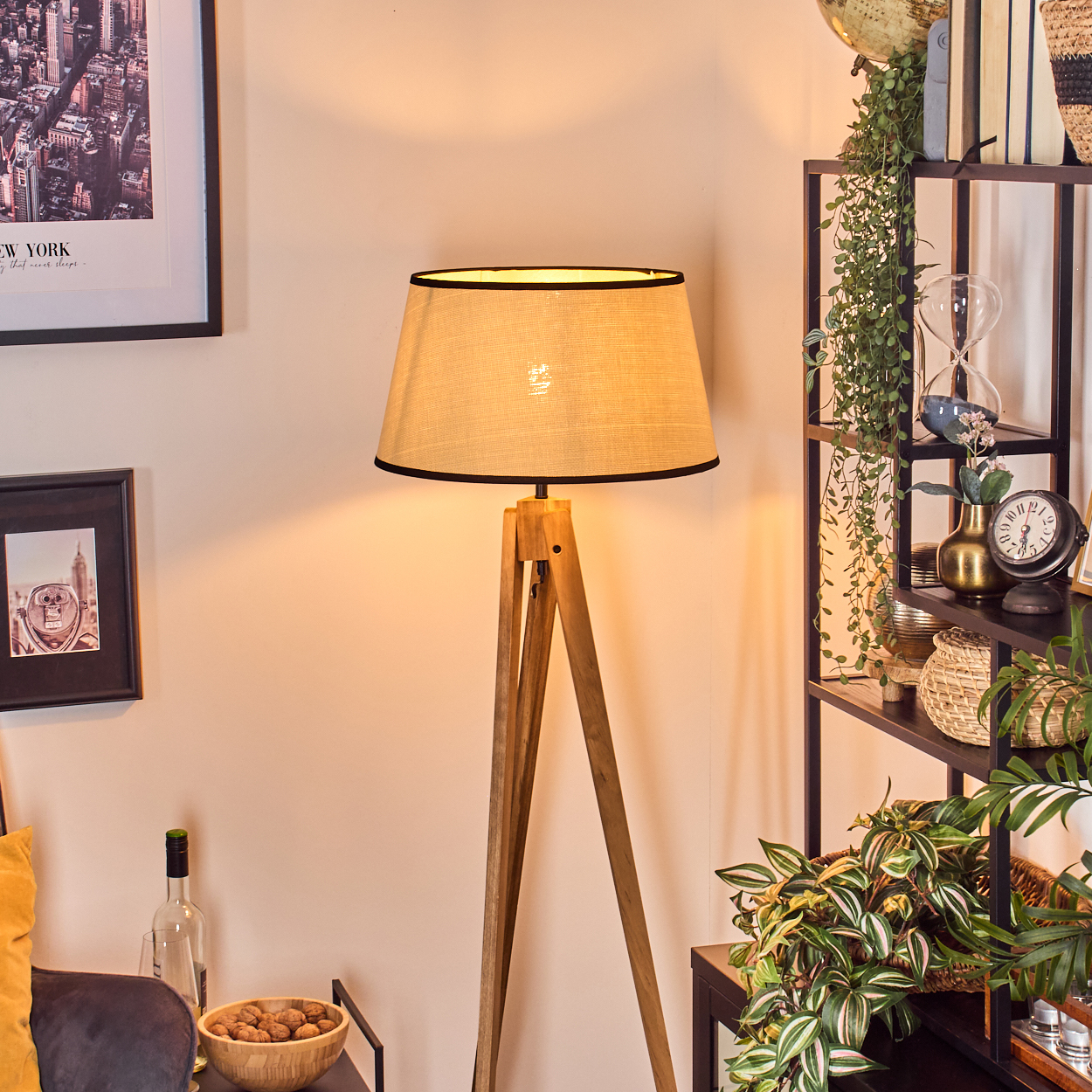 Eine hölzerne Stehlampe mit beigem Schirm steht in einem Raum mit Bücherregal und Bildern.
