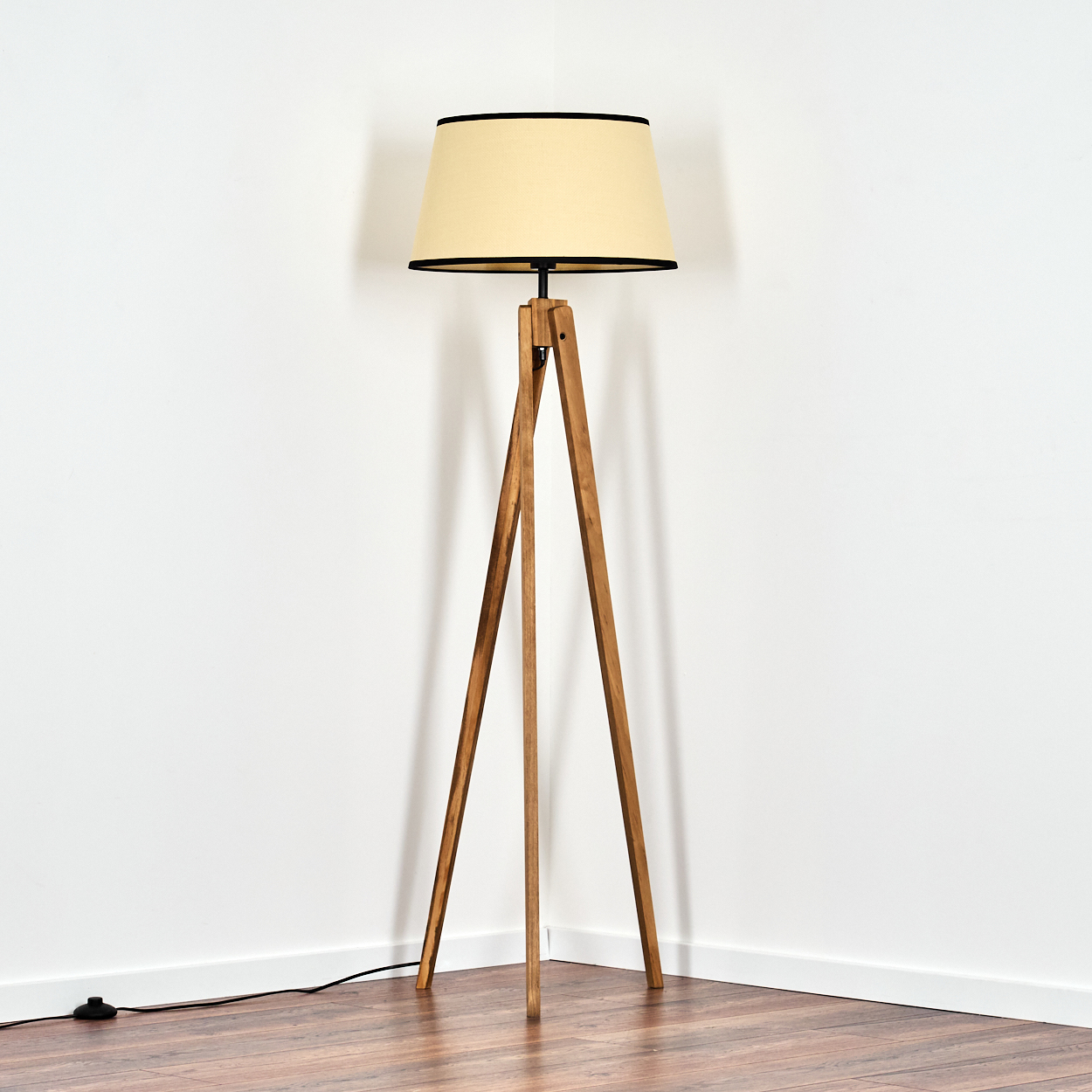 Eine hölzerne Stativ-Stehlampe mit beigem Lampenschirm steht in einer Ecke. Der Boden ist aus Holz, die Wände sind weiß.