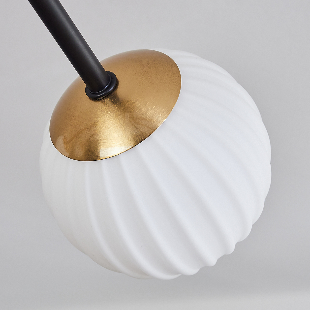 Nahaufnahme einer modernen Leuchte. Merkmale: weißer, geriffelter Glaskugel mit goldener Kappe und schwarzem Arm vor grauem Hintergrund.