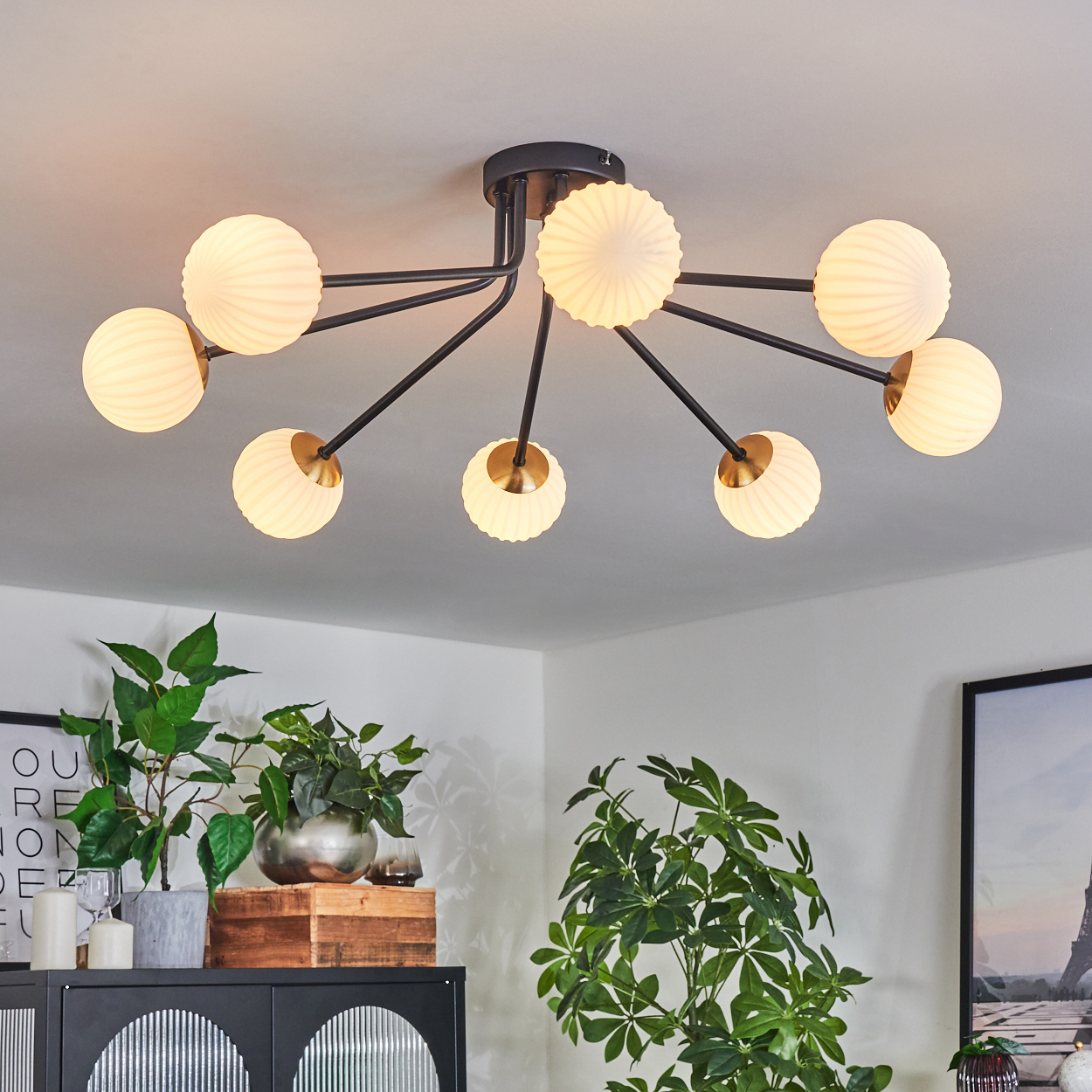 Eine moderne Deckenleuchte mit acht kugelförmigen Lampen. Darunter ein Schrank mit Pflanzen und gerahmten Bildern in einem hellen Raum.