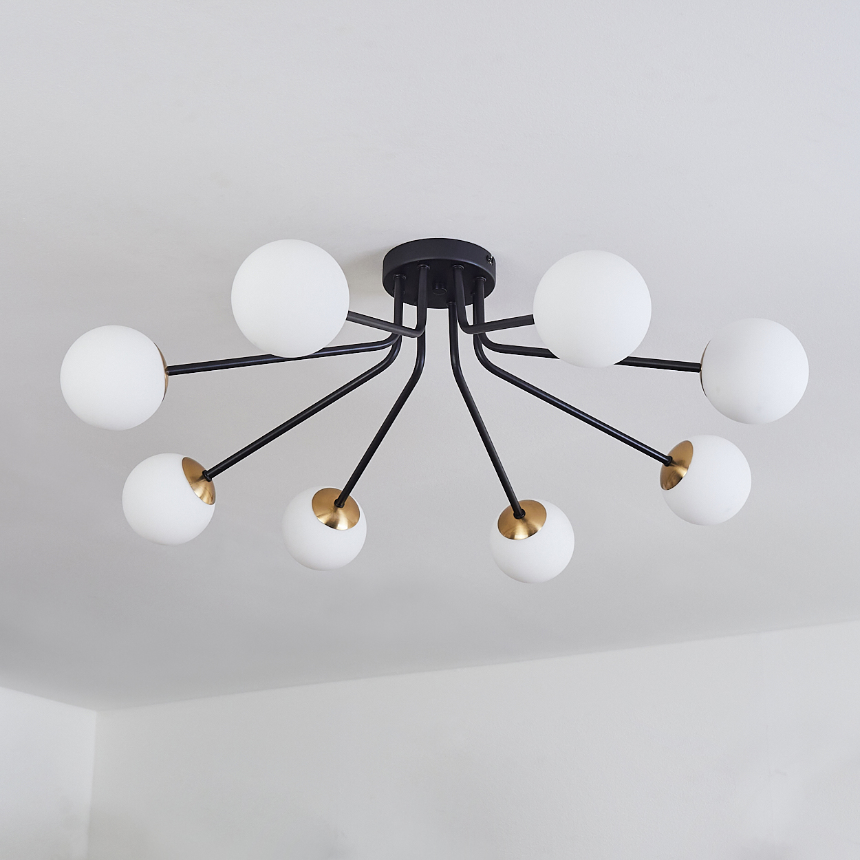 Eine Deckenleuchte mit acht weißen Kugellampen, die an schwarzen, gebogenen Armen befestigt sind, an einer weißen Decke. Einige goldene Akzente.