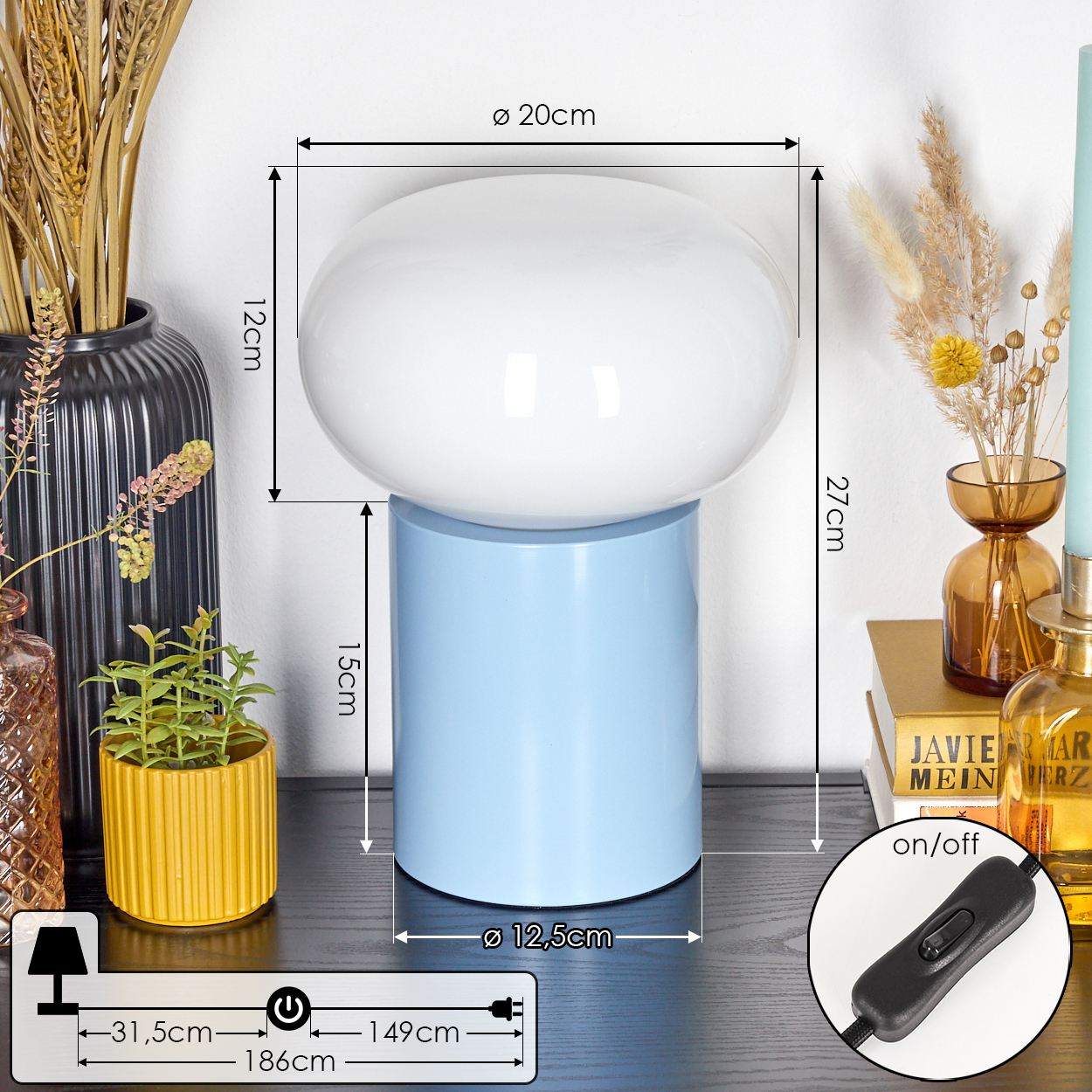 Eine Lampe mit weißem Oberteil und hellblauem Sockel steht auf einem Tisch. Weitere Objekte sind Vasen, Kerzen und eine Pflanze.