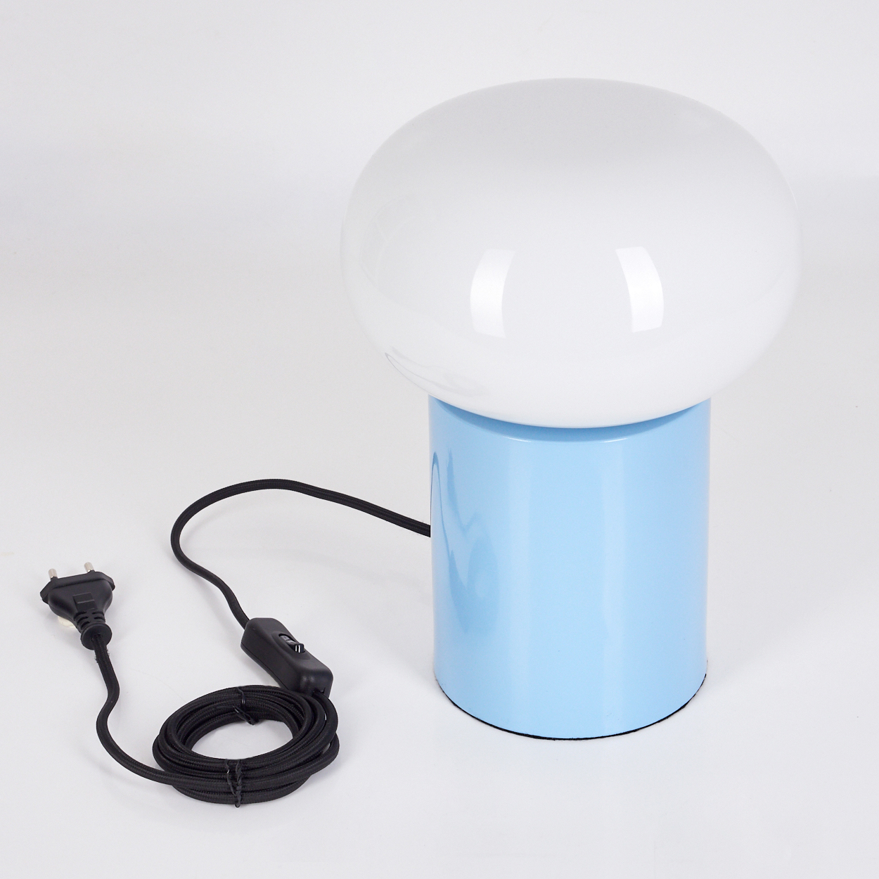 Eine moderne Pilzlampe. Weiße Kuppel auf hellblauem Zylindersockel. Schwarzes Kabel mit Stecker und Schalter. Weißer Hintergrund.