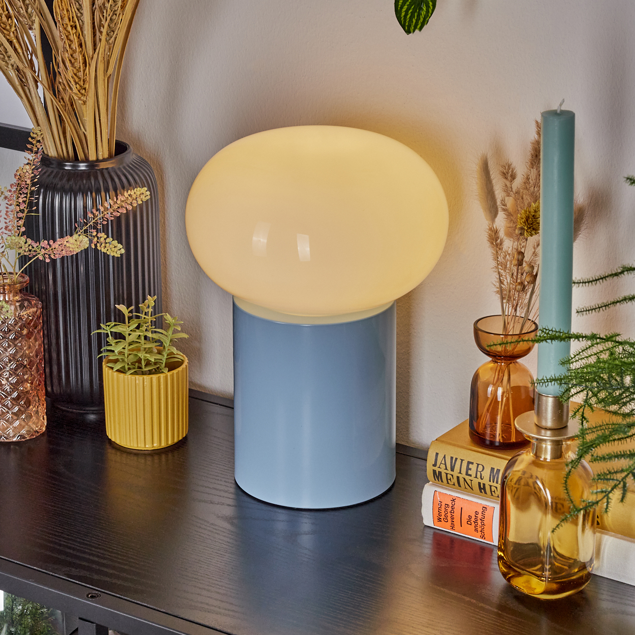 Eine blau-gelbe Lampe steht auf einem dunklen Tisch mit Dekoration. Enthält Vasen, Pflanzen, Bücher und eine Kerze.