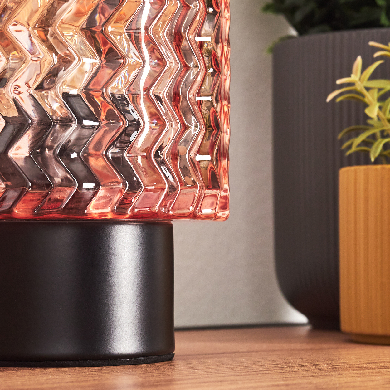 Nahaufnahme einer Glaslampe mit Zickzackmuster. Die Lampe hat einen schwarzen Sockel. Im Hintergrund befinden sich weitere Töpfe mit Pflanzen.
