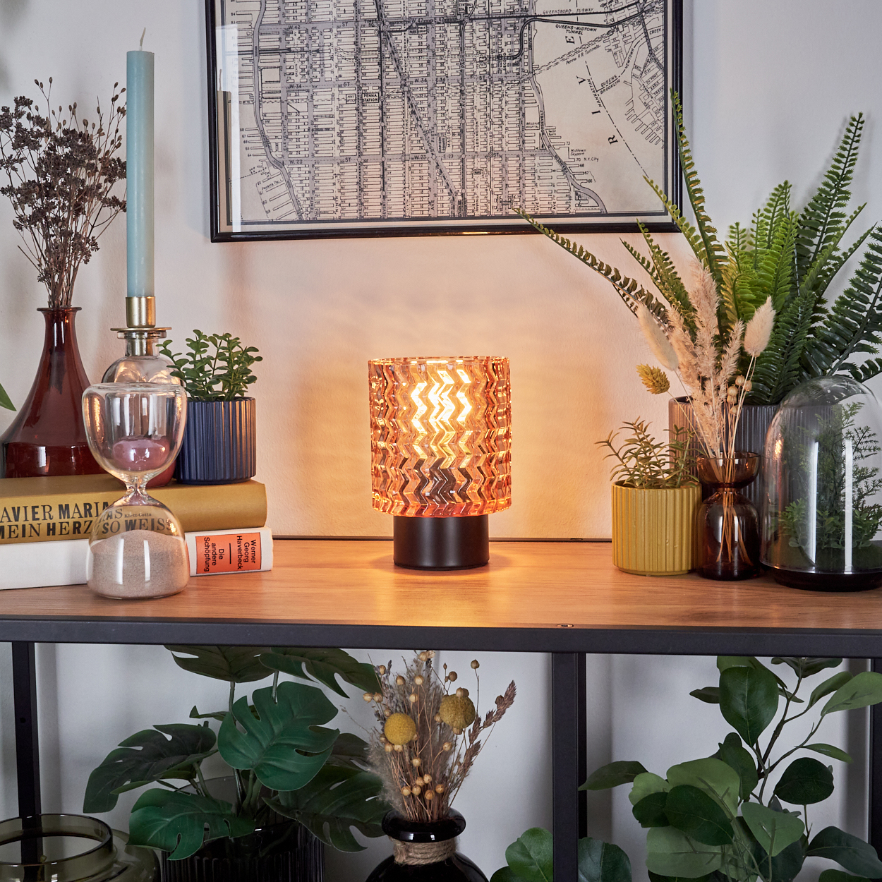 Ein Regal zeigt eine Lampe, Pflanzen und eine Karte. Eine Sanduhr steht neben Büchern. Eine Vase mit Trockenblumen ist ebenfalls vorhanden.