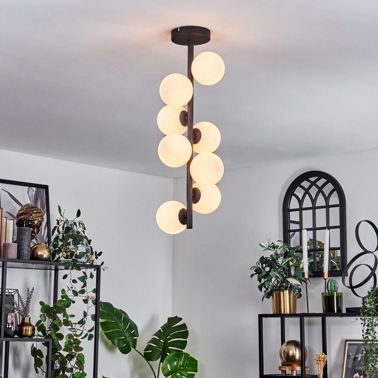 Eine moderne Pendelleuchte mit Kugellichtern hängt von der Decke. Darunter zeigen Regale Pflanzen, Dekoration und einen schwarzen Bogen.