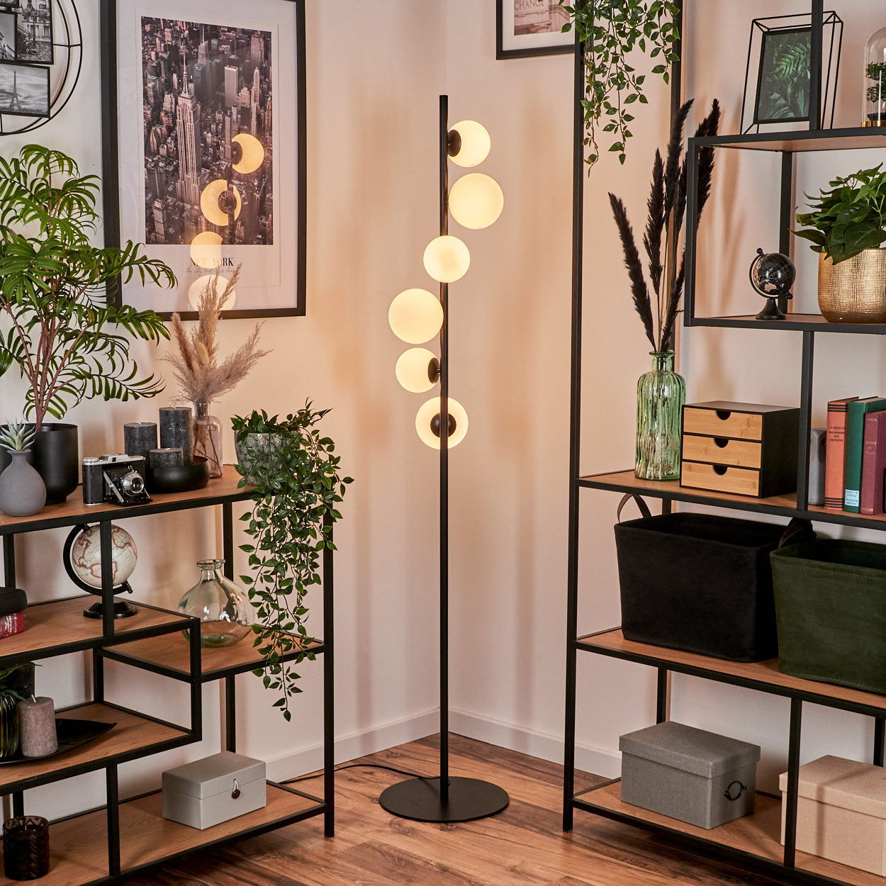 Eine Zimmerecke mit einer modernen Lampe, Regalen mit Dekoration und Pflanzen. Ein Stadtbild hängt an der Wand.