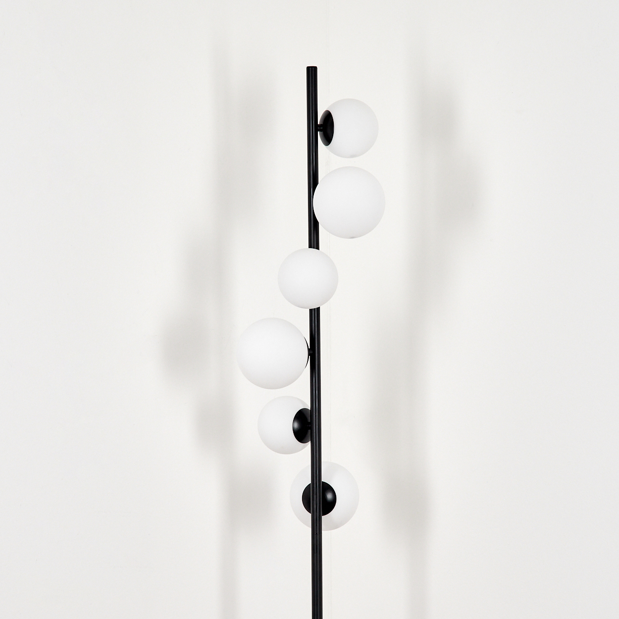 Eine schwarze Metall-Stehlampe mit sechs kugelförmigen, weißen Glasschirmen. Die Lampe steht vor einer schlichten, weißen Wand. Schatten sind sichtbar.