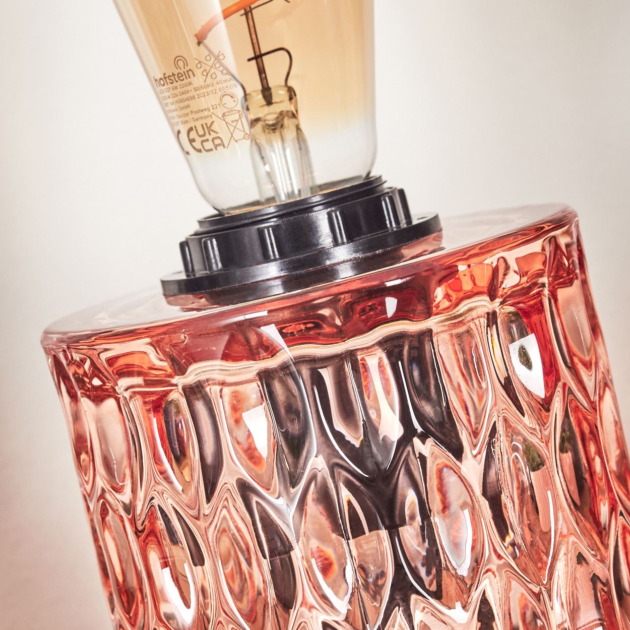 Nahaufnahme einer rosa Glaslampenbasis mit strukturierter Oberfläche, gekrönt von einer schwarzen Halterung und einer Vintage-Glühbirne.