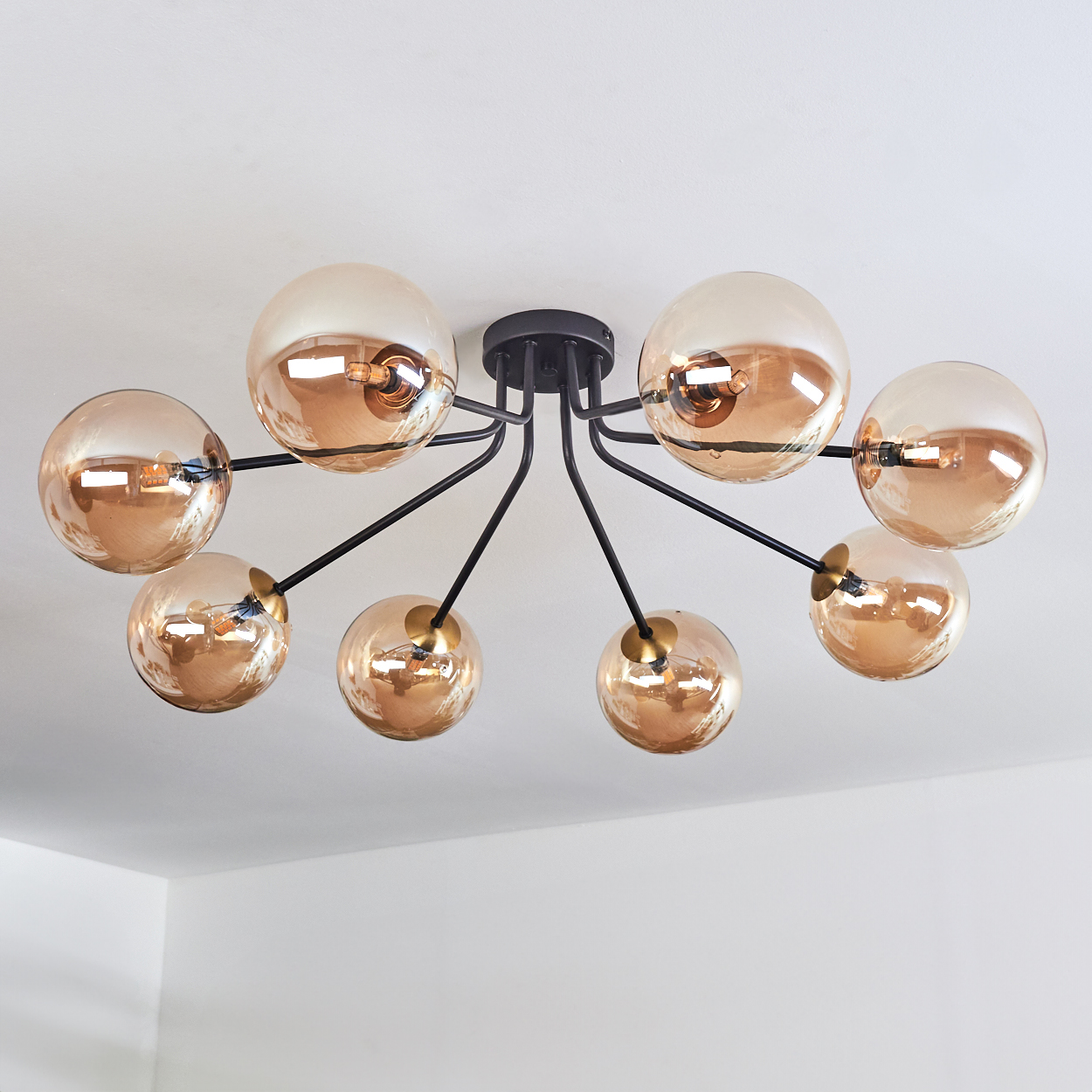 Eine moderne Deckenleuchte mit acht bernsteinfarbenen Glaskugelschirmen, schwarzen Armen und goldenen Akzenten, vor einer weißen Decke.