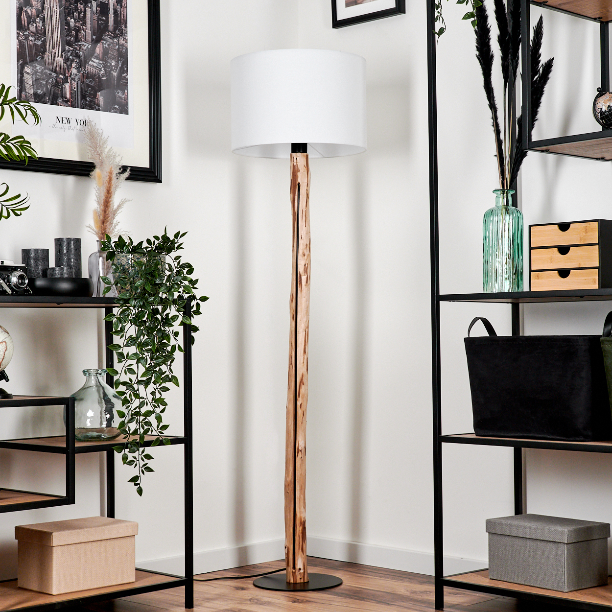 Eine Stehlampe aus Holz mit weißem Lampenschirm steht in einem Raum mit Regalen. Regale halten Dekorationsgegenstände und einen schwarzen Rahmen.