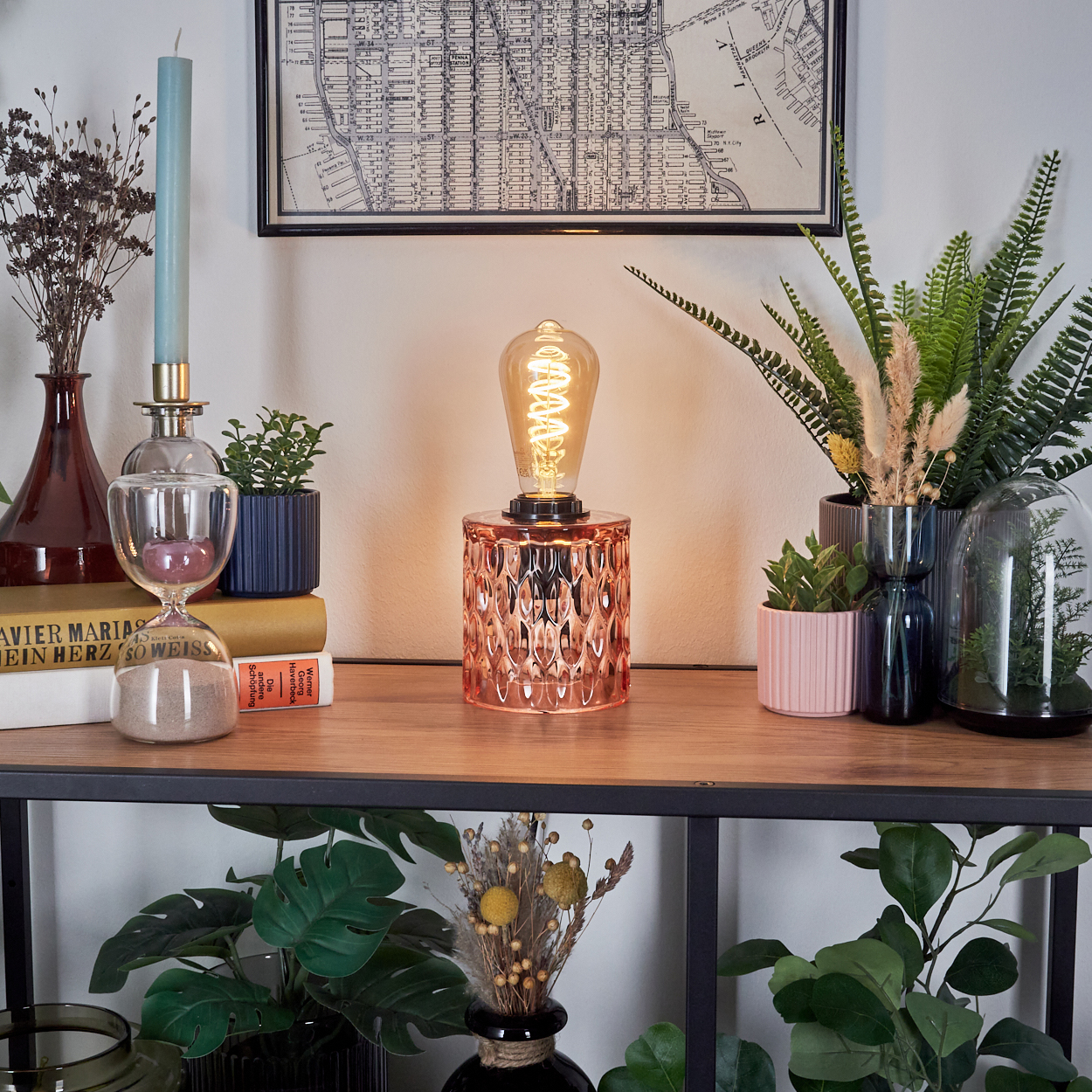 Ein Tisch zeigt eine beleuchtete Lampe, Pflanzen, Bücher und eine Karte. Die Lampe ist der Mittelpunkt, umgeben von Dekorationsartikeln und Grün.