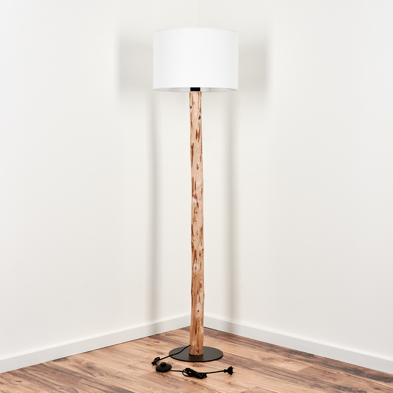 Eine Stehlampe mit Holzstamm und weißem Schirm steht in einer Ecke. Sie hat einen natürlichen Holzstamm und einen schwarzen Sockel.