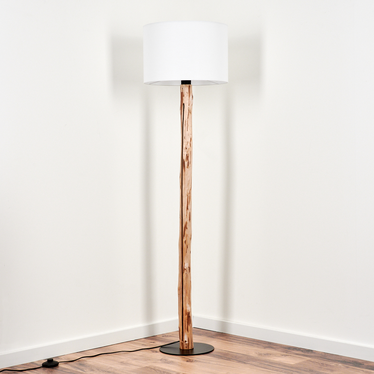 Eine Stehlampe mit weißem Lampenschirm steht in einer Ecke. Die Lampe hat einen Naturholzstamm und einen schwarzen Sockel.