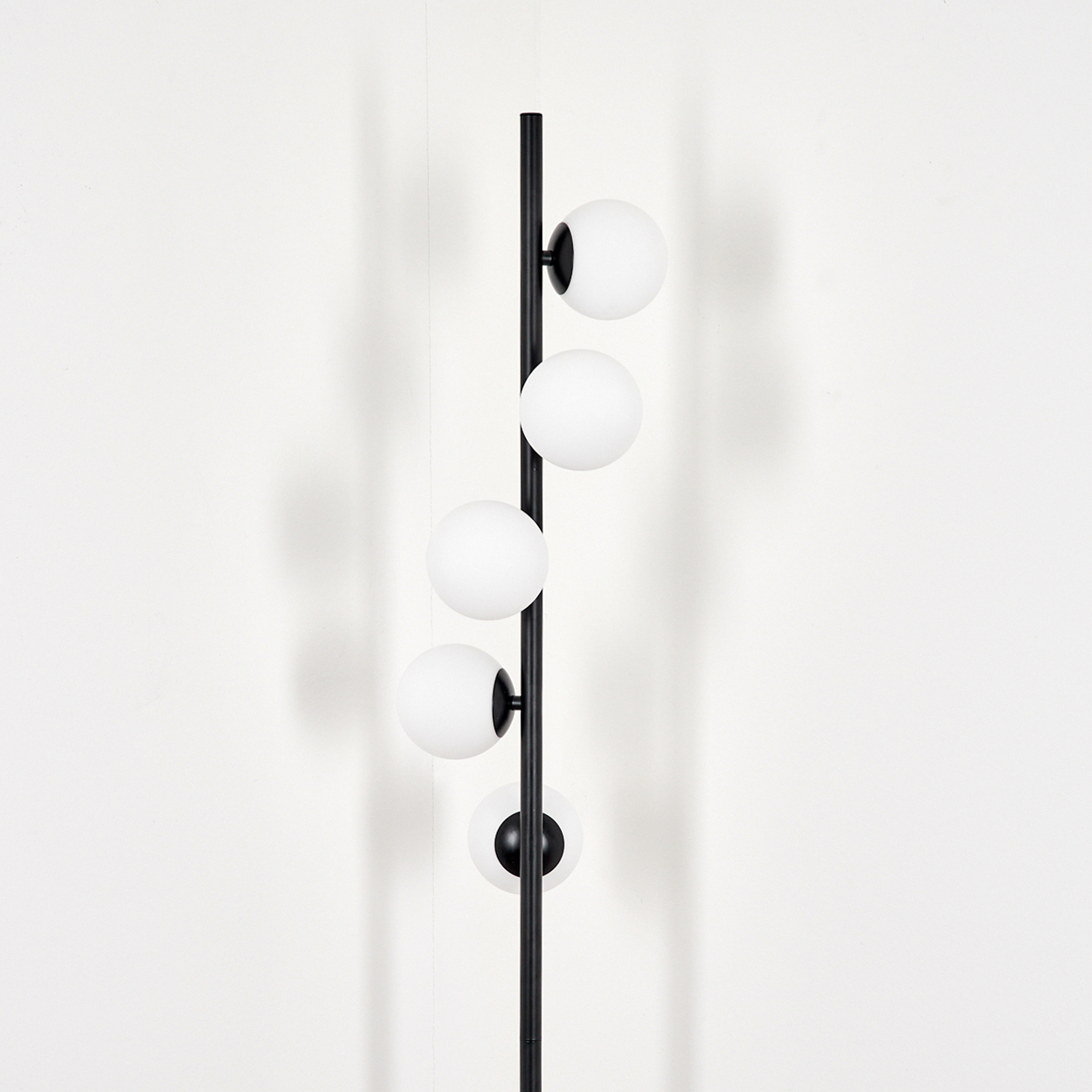 Eine schwarze Stehlampe mit vier weißen Kugelleuchten vor einer weißen Wand. Schatten der Lampe sind sichtbar.