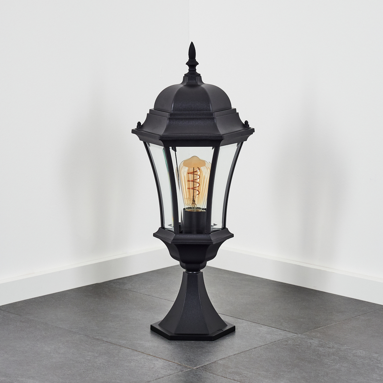 Eine schwarze Außenlampe mit Glasgehäuse und sichtbarer Glühbirne, vor einer weißen Wand und grauem Boden.