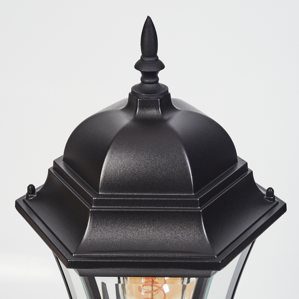 Nahaufnahme einer schwarzen Außenlampe. Sie hat eine dekorative Oberseite mit einem spitzen Aufsatz. Klare Glasscheiben und eine Glühbirne sind sichtbar.