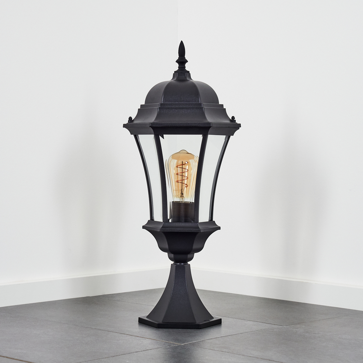Schwarze Außenlampe mit einer leuchtenden Vintage-Glühbirne. Sie steht auf einem grauen Fliesenboden vor einer weißen Wand.