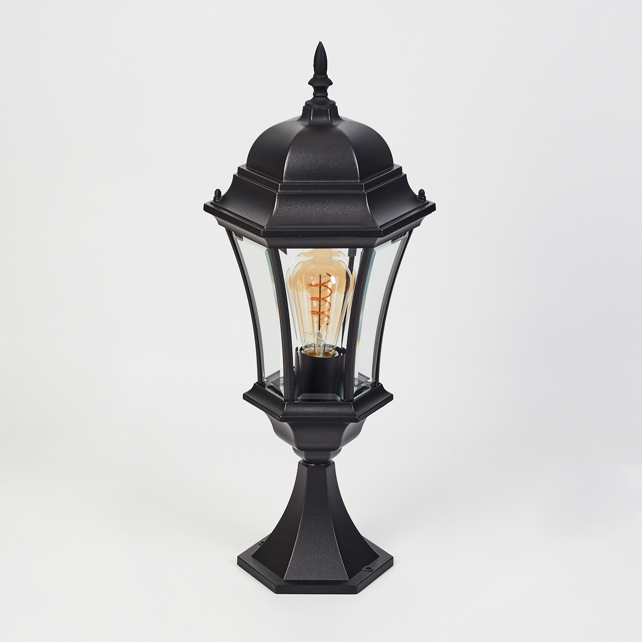 Eine schwarze Außenlampe mit Glaseinfassung und dekorativer Glühbirne, vor weißem Hintergrund.