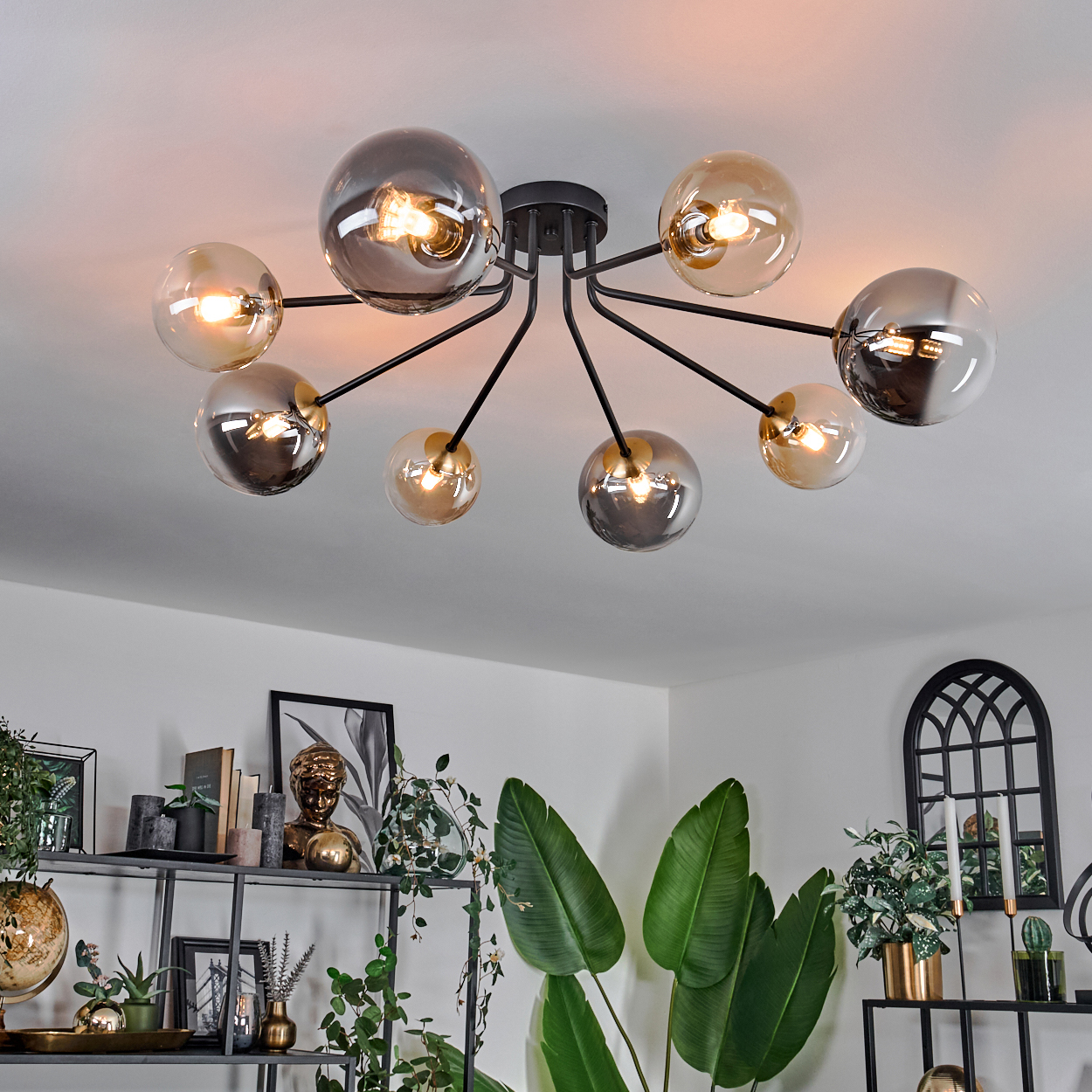 Eine moderne Deckenleuchte mit Glaskugeln. Darunter ein Raum mit Pflanzen, Regalen und Dekoration. Das Licht ist an.