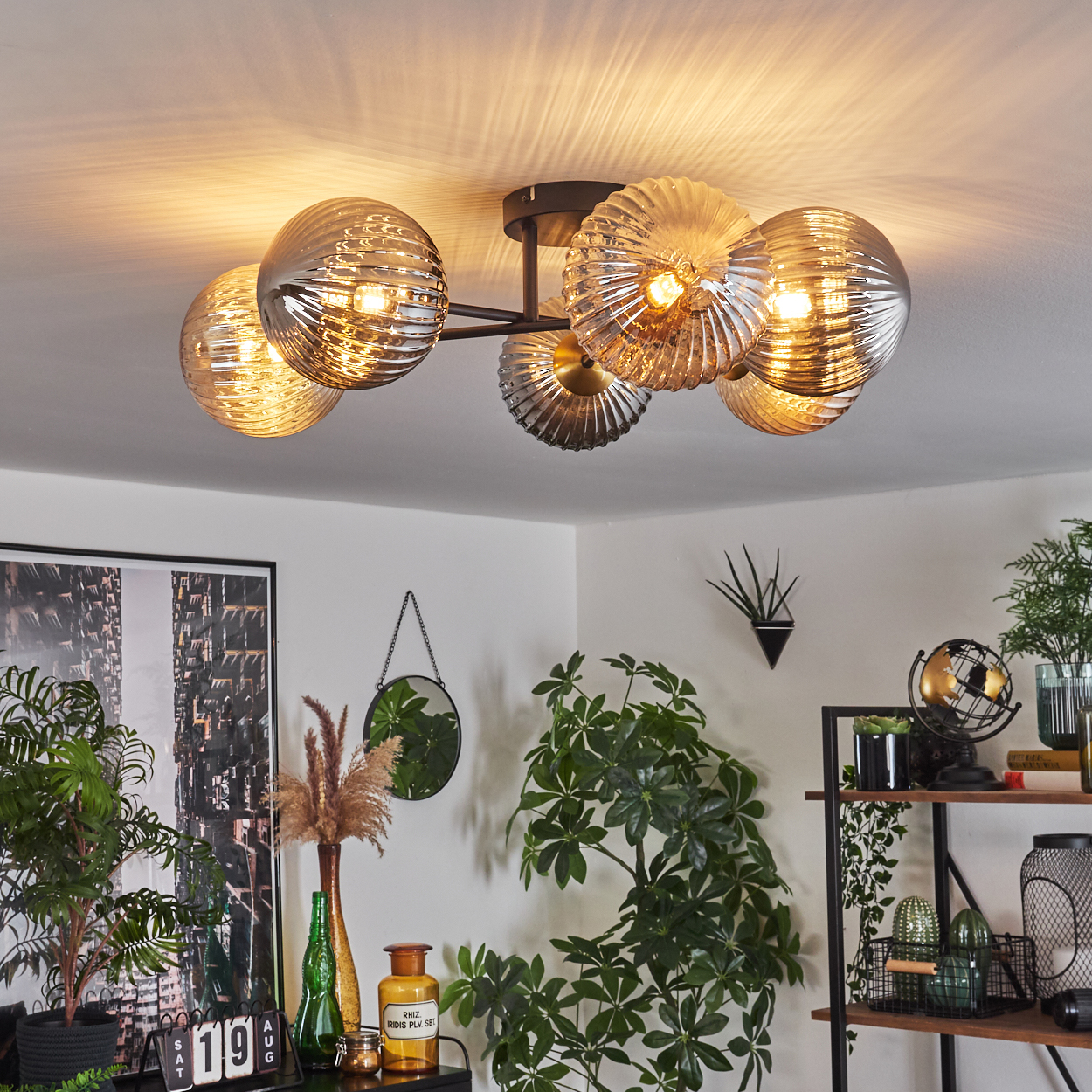Eine Deckenleuchte mit Glaskugeln beleuchtet einen Raum. Pflanzen, ein Spiegel und ein Regal mit Dekoration sind sichtbar.