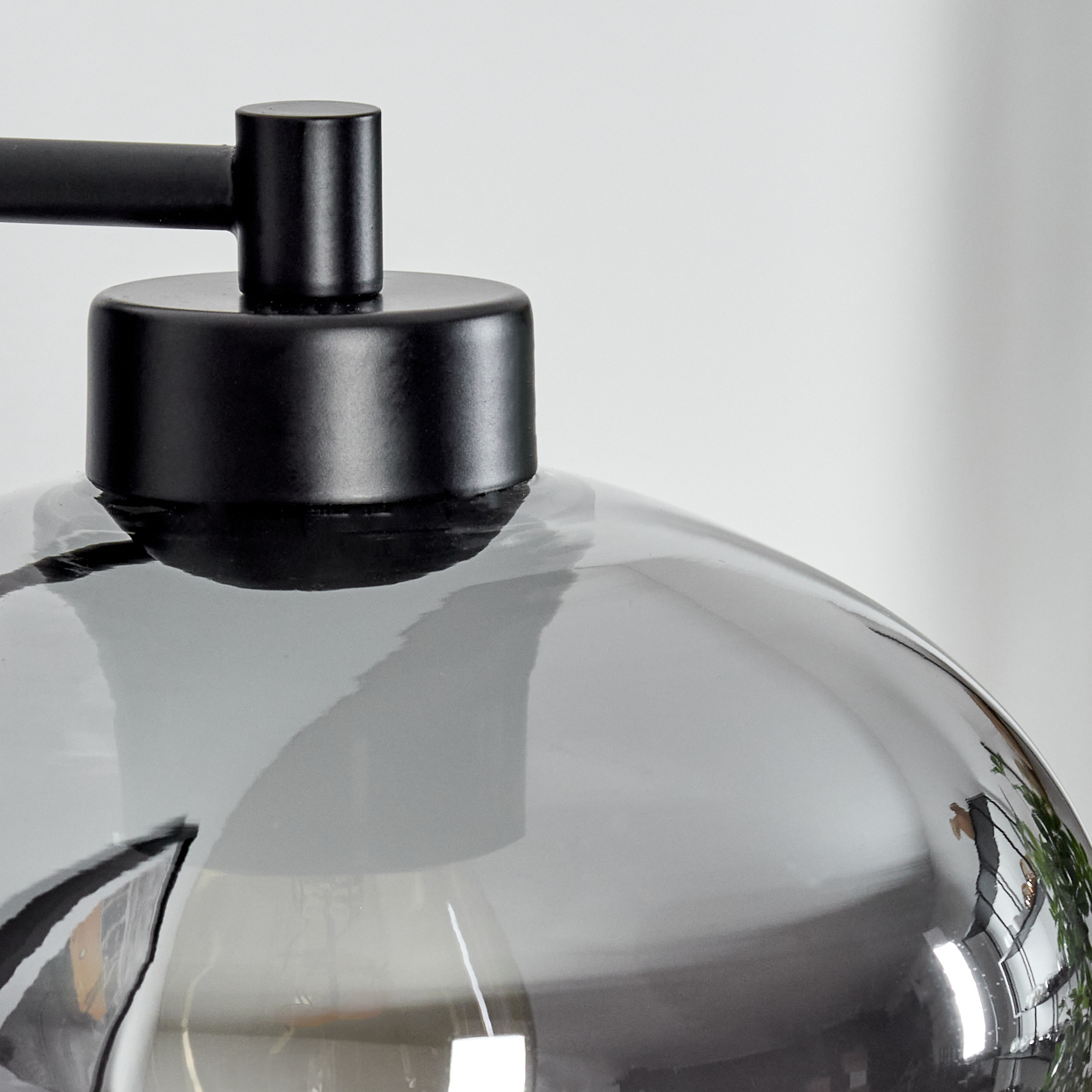 Nahaufnahme einer modernen Lampe. Schwarzer Metallrahmen und -kappe mit rauchigem Glasschirm. Die Reflexion zeigt eine Pflanze.