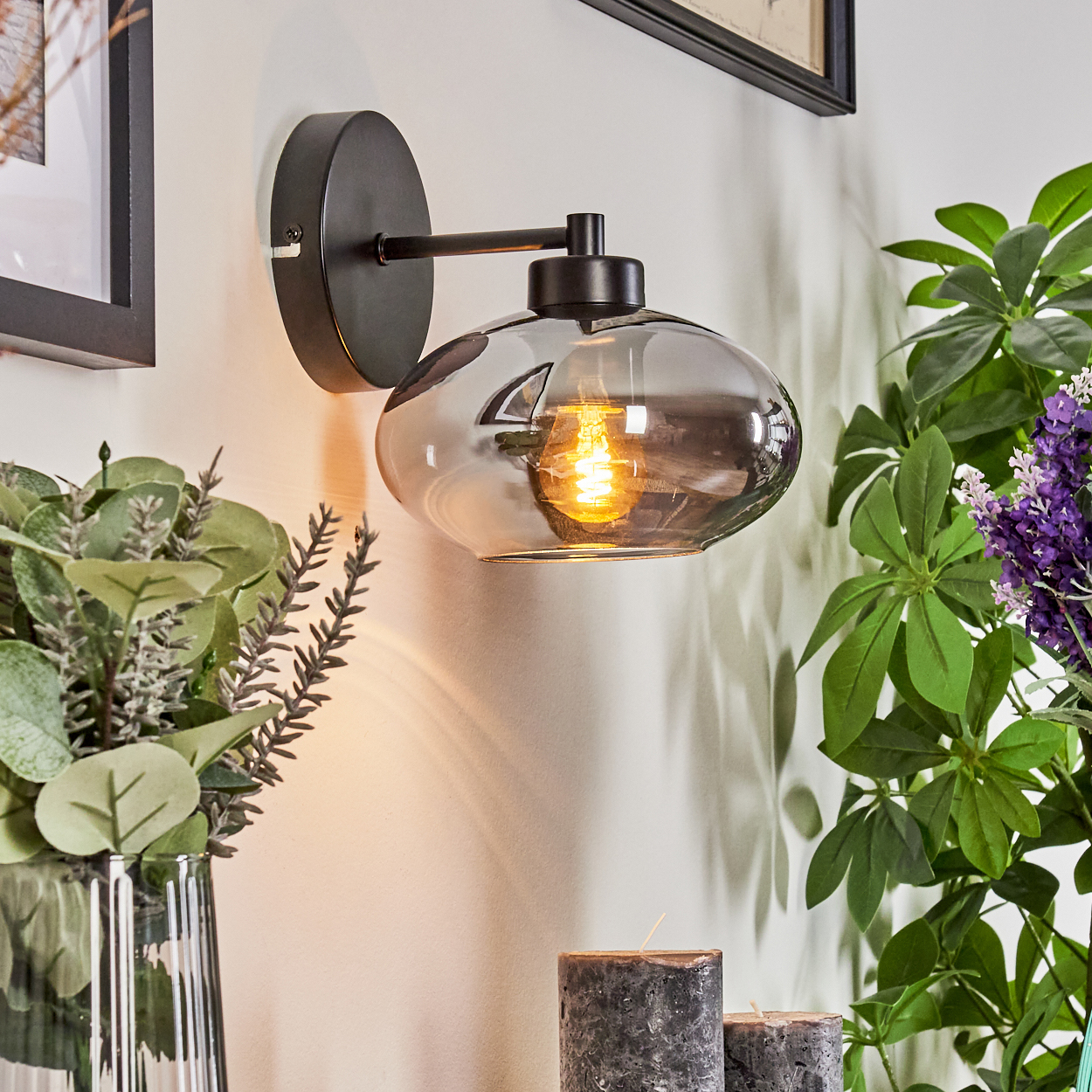Eine Wandlampe mit rauchfarbenem Glasschirm leuchtet. Daneben stehen grüne Pflanzen und einige Kerzen. Der Hintergrund ist eine weiße Wand.