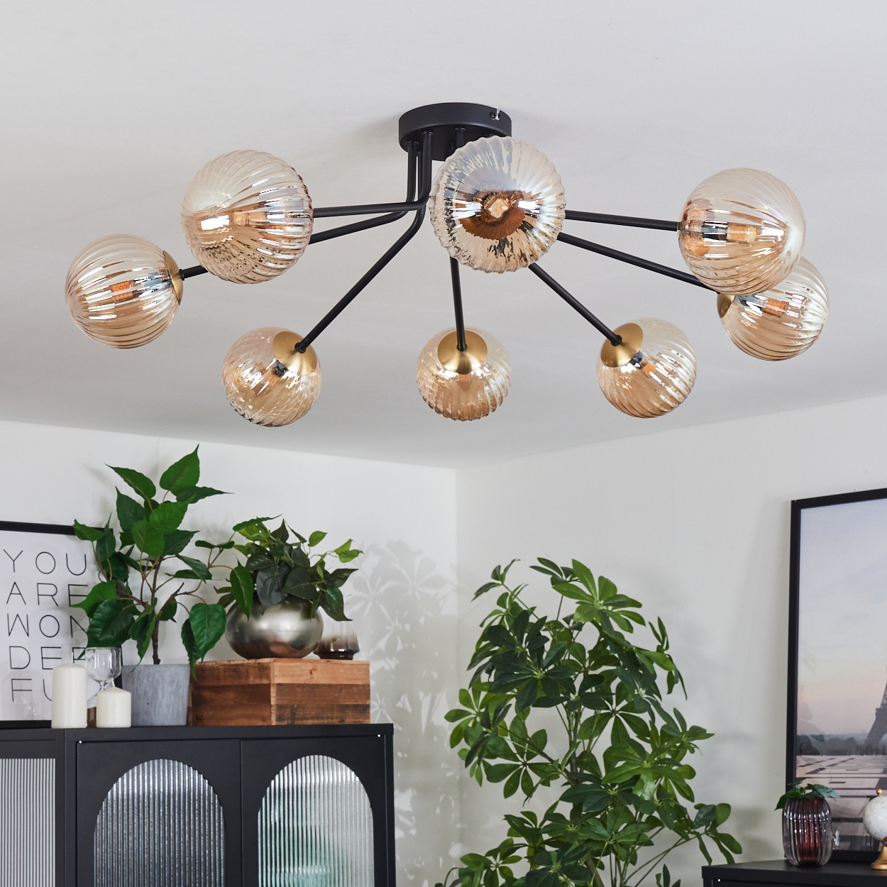 Deckenlampe mit Glaskugeln, schwarzen Armen. Pflanzen, Kerzen und ein schwarzer Schrank im Vordergrund. Weiße Wände.