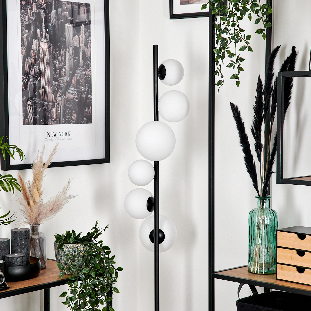 Eine Zimmerecke mit Lampe, New York-Druck, Pflanzen und Dekorationsartikeln auf Regalen. Die Lampe hat weiße Kugeln.